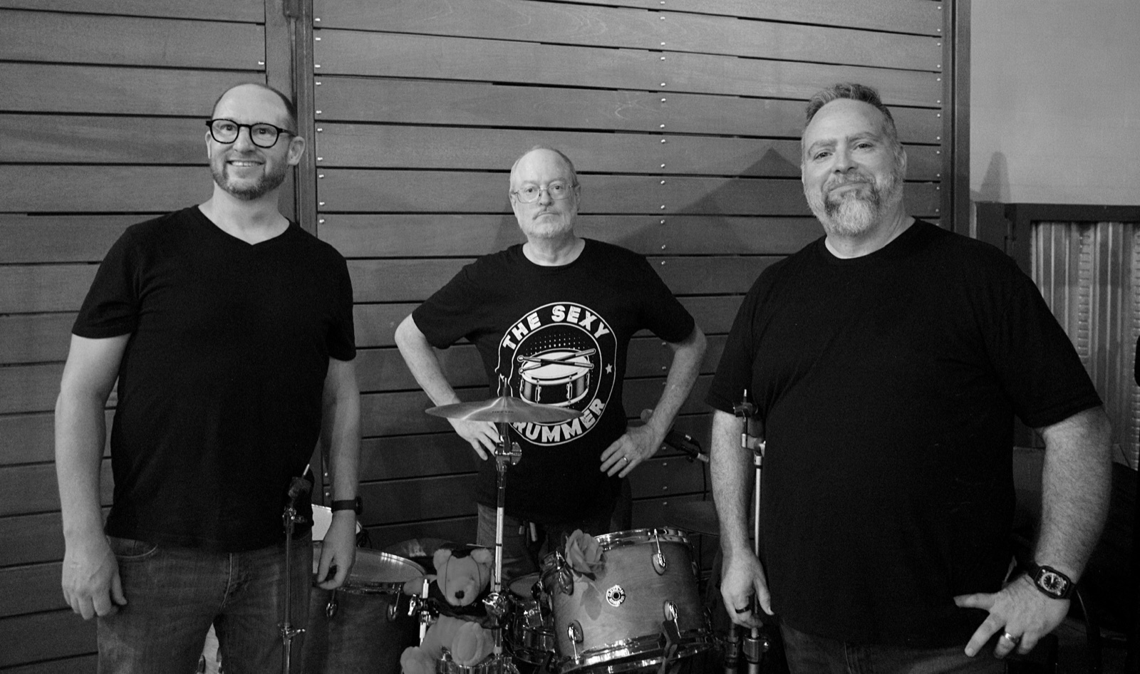 The three members of BGM posing in front of a wood-slat wall behind a drum kit